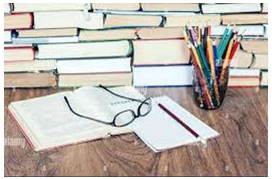 A photo of a desk with piles of books, an open notebook with reading glasses, and a pot of coloured pencils.
